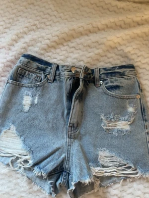 Blå slitna jeansshorts med hög midja - Säljer ett par blå denimshorts med hög midja och råa, slitna detaljer. Shortsen har klassisk femficksdesign, bälteshällor och knappgylf.