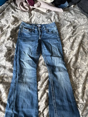 Blå bootcut jeans med broderade fickor - Säljer ett par blå bootcut jeans med snygga broderade detaljer och knappar på bakfickorna. Jeansen har klassisk femficksmodell och är tillverkade i denim med lätt slitning framtill och baktill. Perfekta för dig som gillar en avslappnad men trendig stil.
