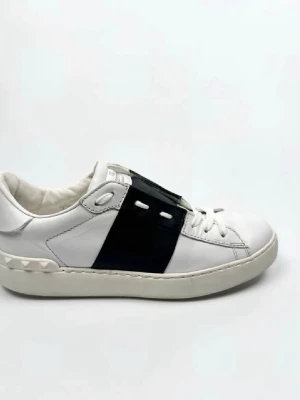 Valentino vita sneakers med svart detalj - Snygga vita sneakers från Valentino med en bred svart läderdetalj över snörningen och ikoniska nitar på hälen. Skorna har en klassisk låg profil och är tillverkade i slätt läder med vita snören och sula.
