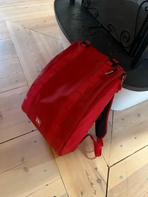 Röd ryggsäck från Douchebags 20L - Snygg röd ryggsäck från Douchebags med stilren design och praktiska dragkedjor. Väskan har vadderade axelband och baksida för extra komfort samt flera fack för smart förvaring. Perfekt för dig som vill ha en cool och funktionell väska till skolan eller stan.