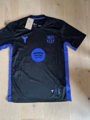 Svart FC Barcelona matchtröja Nike - Snygg svart FC Barcelona matchtröja från Nike med blå detaljer och tryck. Tröjan har klubbmärke, Spotify-logga och nummer 10 med Lamine Yamal på ryggen. Tillverkad i lätt och ventilerande funktionsmaterial, perfekt för fotboll eller träning.