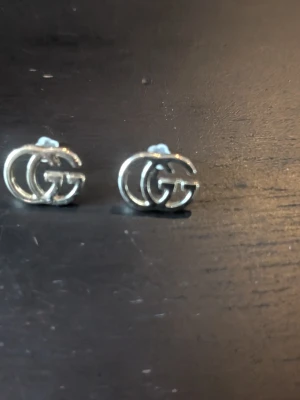 Guldiga GG örhängen  - Säljer ett par stilrena örhängen i silverfärgad metall. Örhängena har den klassiska dubbla G-loggan och är små och diskreta, perfekta för dig som gillar exklusiva accessoarer med ikonisk design.