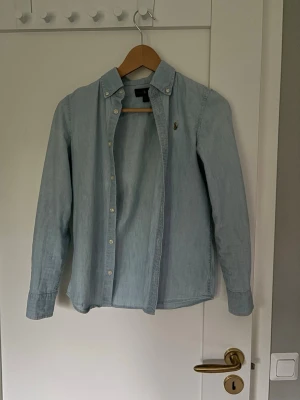 Ljusblå skjorta från Ralph Lauren - Säljer en klassisk ljusblå skjorta från Ralph Lauren med button-down krage och broderad logga på bröstet. Skjortan har långa ärmar, vita knappar och är tillverkad i mjuk bomull. 
