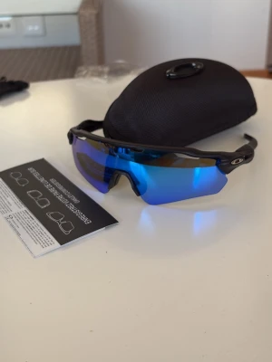 Oakley radar  - Säljer ett par sportiga solglasögon från Oakley med spegelblå linser och svart båge. De har en modern, svepande form som passar perfekt för utomhusaktiviteter. Kommer med ett hårt fodral och instruktionsblad.