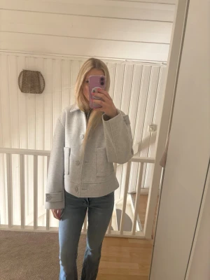 Ljusgrå kort jacka med knappar - Snygg ljusgrå jacka med rak passform och stora fickor framtill. Jackan har knappar längs framsidan och vid ärmsluten, samt en croppad design som ger en modern look. Perfekt för lager-på-lager-stil.