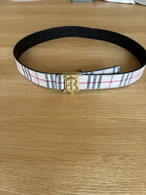 Dubbelsidig Burberry bälte med guldspänne - Snyggt bälte från Burberry med klassiskt rutigt mönster i vitt, grått, svart och rosa. Bältet har ett stilrent guldigt B-spänne framtill och är tillverkat i skinn. Perfekt accessoar för att lyfta din outfit.             Pris kan diskuteras.