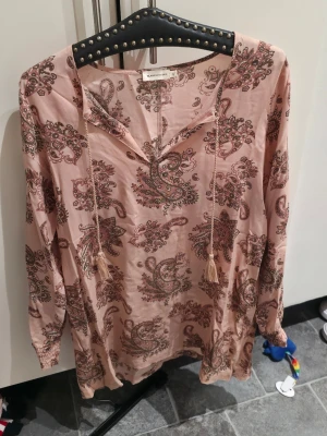 Mönstrad blus från Angelology - Ljusrosa blus från Angelology med paisleymönster i brunt och grönt. Blusen har långa ärmar, v-ringning med knytband och tofsar. Tillverkad i ett mjukt, tunt tyg som ger en avslappnad och bohemisk känsla.