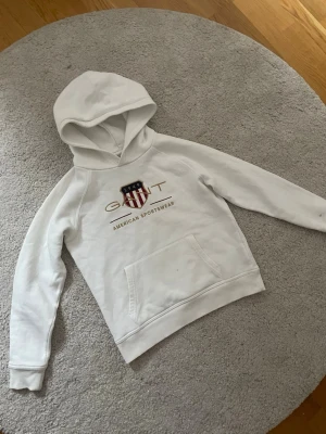 Vit hoodie från Gant med tryck - Snygg vit hoodie från Gant med huva och magficka. På bröstet finns ett broderat Gant-emblem i rött, blått och guld samt texten 'American Sportswear'. Hoodien har klassisk passform och är tillverkad i mjuk bomullsblandning. Jätte bra skick det ändå är två små små fläckar som visas på bilden men syns knappt och går nog bort om man skrubbar bort det 