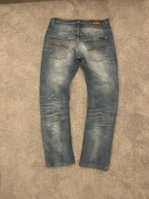 Nudie Jeans selvage  - Säljer ett par blå jeans från Nudie Jeans i storlek 32/32 med slitningar på knäna. Jeansen har klassisk femficksdesign, raka ben och ljus tvätt med snygga detaljer på bakfickorna. Perfekta för en avslappnad och trendig stil.