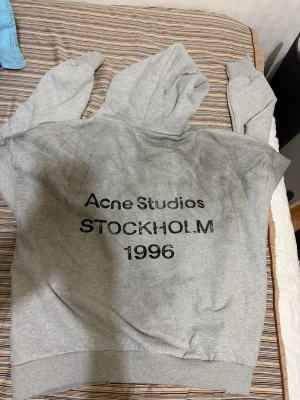 Grå hoodie från Acne Studios - Snygg grå hoodie från Acne Studios med tryck 'Acne Studios STOCKHOLM 1996' på framsidan. Klassisk passform med huva och ribbade muddar. Perfekt för en avslappnad streetwear-look. Materialet är mjukt och skönt bomullstyg.