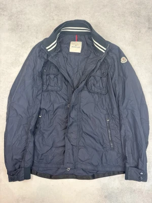 Marinblå vindjacka från Moncler - Snygg marinblå vindjacka från Moncler med två bröstfickor med lock, två sidofickor med dragkedja och Moncler-logga på ärmen. Jackan har ståkrage med vita ränder och dragkedja framtill. Tillverkad i lätt, glansigt material.