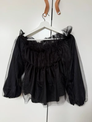 Svart offshoulder meshblus - Trendig svart blus i mesh med offshoulder-modell och puffiga, långa ärmar. Blusen har volangdetaljer vid halsringningen och är tillverkad i ett lätt, transparent syntetmaterial som ger en luftig och edgy look. Perfekt till fest!!