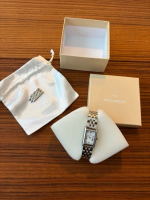 Mockberg silver klocka - Stilren klocka från Mockberg med rektangulärt vitt ur och romerska siffror. Armbandet är i silverfärgad metall med klassisk länkdesign. Kommer med originalask och extra länk. Perfekt accessoar för en clean och elegant look.
