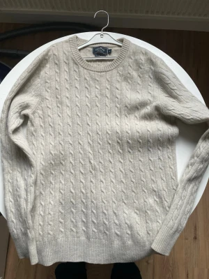 East West Beige kabelstickad tröja - East West Snygg beige kabelstickad tröja med klassiskt mönster och ribbade muddar. Perfekt för lager-på-lager och passar till många olika stilar. Mjuk och skön känsla, rund halsringning och lång ärm.  Ullkvalite