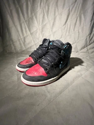 Nike Air Jordan 1 High svart/röd/blå - Snygga Nike Air Jordan 1 High sneakers i svart läder med röda detaljer på tån och blå detaljer vid häl och swoosh. Klassisk siluett med högt skaft, svarta skosnören och vit sula. Perfekta för dig som gillar streetwear och vill sticka ut.skorna är lite slitna på båda insidorna
