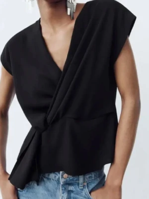 Svart omlottopp med knyt - Stilren svart omlottopp med knyt i sidan och v-ringad hals. Toppen har korta ärmar och en lös passform som ger en avslappnad men trendig look. Perfekt att matcha med jeans eller kjol för en cool vibe.