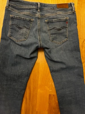 Blå jeans från Replay - Säljer ett par klassiska blå jeans från Nudie Jeans med smal passform. Jeansen har snygga bakfickor med vågigt broderi och en liten logga på ena fickan. Tillverkade i denim med en lätt tvättad look. Perfekta för en avslappnad stil. L30