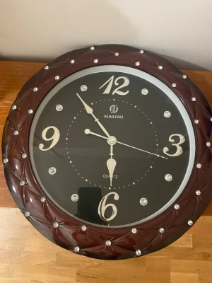 Rund väggklocka  - Säljer en rund väggklocka med mörkbrunt ram i plast och dekorativa stenar runt kanten. Urtavlan är svart med stora silvriga siffror och detaljer. Klockan drivs med batteri och har tydliga visare.