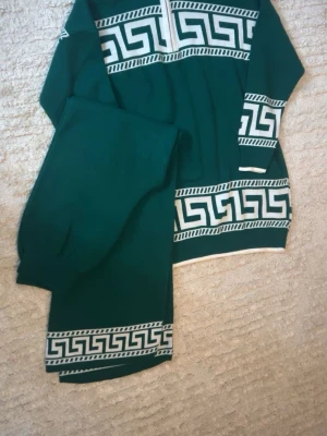 Grönt loungewear-set med mönster - Snyggt grönt loungewear-set med vita grekiska mönster längs ärmar, midja och benslut. Tröjan har halv dragkedja framtill och lång ärm. Mjukt material som känns skönt mot huden, perfekt för chill dagar hemma.