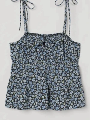 Blommig linnetopp med knytband - Söt linnetopp med blått och vitt blommönster på svart botten. Toppen har smala axelband med knyt och en liten rosett framtill. Perfekt för varma dagar och enkel att styla med jeans eller kjol.