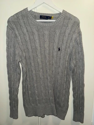 Grå kabelstickad Polo Ralph Lauren tröja - Klassisk grå kabelstickad tröja från Polo Ralph Lauren med rund halsringning och den ikoniska mörkblå logotypen broderad på bröstet. Tröjan har långa ärmar och ribbade muddar vid ärmslut och nederkant. Perfekt för lager på lager och stilren vardagslook.