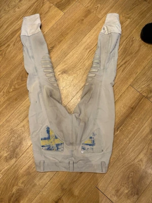 Ljusgrå ridbyxor med svensk flagga från animo - Snygga ljusgrå ridbyxor från animo med detaljer i form av svenska flaggan på bakfickorna och förstärkta partier på insidan av benen. Byxorna har ribbade knäpartier och mudd vid benslut för extra komfort.