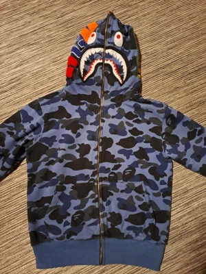 BAPE blå camo shark hoodie - Säljer en blå camo zip hoodie från BAPE med ikonisk shark-design på huvan och färgglada detaljer. Hoodien har dragkedja hela vägen, kängurufickor och ribbade muddar. Perfekt för dig som gillar streetwear och vill sticka ut.