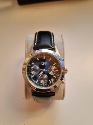 Svart GFF klocka med läderband - Stilren klocka från GFF med svart urtavla och flera små urtavlor. Boetten är i silverfärgat rostfritt stål och armbandet är i äkta svart läder med vita sömmar. Klockan är vattentålig upp till 30 meter och har klassisk rund form.