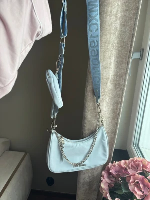 Ljusblå handväska från Steve Madden - Trendig ljusblå handväska från Steve Madden med bred axelrem i textil och silverfärgade detaljer. Väskan har en avtagbar kedja och en liten matchande miniväska som hänger på remmen. Perfekt för dig som gillar streetstyle och snygga accessoarer.