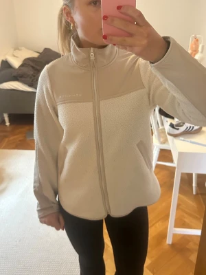 Vit fleecejacka från Röhnisch - Säljer en vit fleecejacka från Röhnisch med hög krage och hel dragkedja framtill. Jackan har två sidofickor och beige detaljer vid axlar och krage. Perfekt för skidresor och afterski!!🥰🥰 andvönd i en vecka förra året och i mkt bra skick🫶🫶