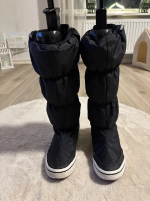Svarta moon boots från Adidas - Säljer ett par svarta moon boots från Adidas med klassiska tre ränder baktill. Bootsens ovandel är i vadderat syntetmaterial och sulan är vit med svart kant. Perfekta för kalla dagar när du vill ha både stil och värme.