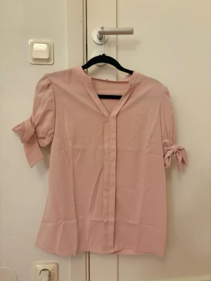 Ljusrosa blus med knytärmar - Söt ljusrosa blus med V-ringning och dekorativa knytband på ärmarna. 