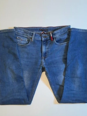 Blå jeans från Tommy Hilfiger - Säljer ett par klassiska blå jeans från Tommy Hilfiger med raka ben och fem fickor. Jeansen har snygga detaljer som Hilfiger-logga på fickan och bak, samt en röd etikett i linningen. Tillverkade i mjukt denimtyg som sitter skönt. Storleken är 176 men passar S. Tveka inte att höra ac er vid frågor/funderingar.