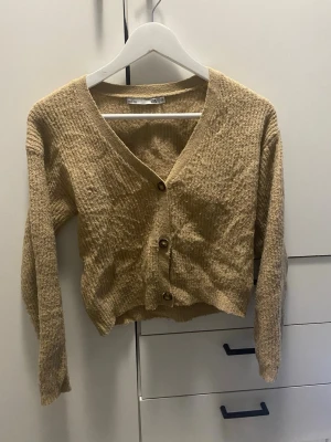 Beige kofta från 157 med knappar - Mysig beige kofta från 157 med v-ringning och tre bruna knappar framtill. Koftan har en croppad passform och är tillverkad i ett mjukt, strukturerat material. Perfekt att slänga över en topp för en avslappnad look.