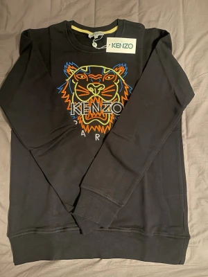 Svart stickad Kenzo tröja tiger - Svart stickad tröja från Kenzo med ikoniskt broderat tigerhuvud i färgglada toner på bröstet. Rund halsringning med gul detalj på insidan och långa ärmar. Perfekt för dig som vill ha en statement piece i garderoben.