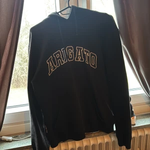 Svart Arigato hoodie med tryck - Säljer en svart hoodie från Arigato med stort vitt tryck över bröstet. Klassisk passform med huva och dragsnören, samt ribbade muddar vid ärmar och midja. Perfekt för dig som gillar streetwear och enkel stil.
