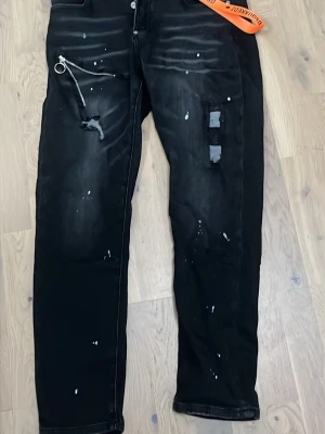 Svarta Dsquared2 jeans storlek 32 (48) - Säljer ett par svarta jeans från Dsquared2 med slitningar, vita färgstänk och coola detaljer som dragkedja och orange DSQUARED2-nyckelband. Jeansen har en modern passform och patchade partier på ena benet. Perfekta för dig som gillar streetstyle.