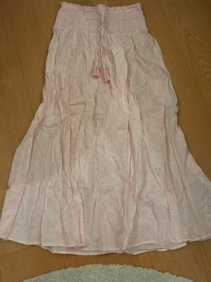 Ljusrosa långkjol med snörning - Söt ljusrosa långkjol med smock i midjan och snörning med tofsar. Kjolen har ett diskret mönster och är tillverkad i ett lätt och luftigt tyg, perfekt för varmare dagar. Jättegullig på sommaren och jag säljer eftersom den är för liten! Skulle säga att den passar en xs. Nästan aldrig använd!