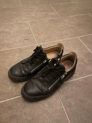 Svarta sneakers med dragkedjor - Säljer ett par svarta sneakers från Giuseppe Zanotti med coola dragkedjor på sidorna och svarta skosnören. Skorna är låga och har en stilren design i läder med bruna detaljer på insidan. Perfekta för dig som gillar streetstyle och exklusiva märken. Box m.m följer med.