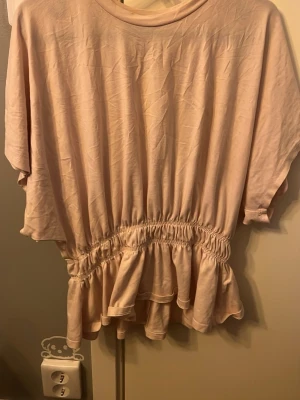 Ljusrosa blus med volang - Säljer en ljusrosa blus i mjukt bomullstyg. Toppen har korta ärmar, rund halsringning och en bred resår i midjan som ger en snygg volangkant nedtill. Perfekt för dig som gillar en feminin och avslappnad stil.