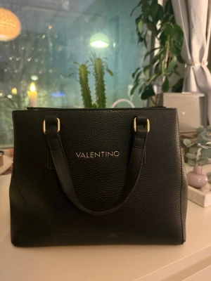 Svart handväska från Valentino - Snygg svart handväska från Valentino i strukturerat konstläder med guldfärgade metalldetaljer. Väskan har två rejäla handtag och en stilren, fyrkantig form. Perfekt för dig som gillar klassisk och elegant design.