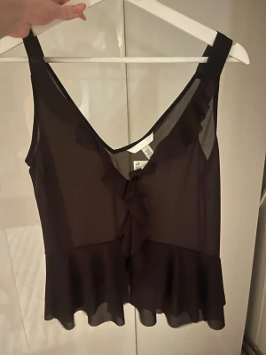 Brun topp - Snygg svart ärmlös topp i transparent material med volangdetaljer framtill och v-ringning. Toppen har en luftig och flowy passform, perfekt för att styla lager på lager. Passar dig som gillar en chic och trendig look.