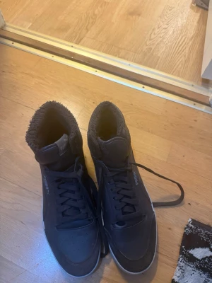 Svarta Puma sneakers med foder - Snygga svarta sneakers från Puma med högt skaft och mjukt foder på insidan. Skorna har klassisk snörning, diskret Puma-logga på sidan och en vit sula som kontrast. Perfekta för dig som vill ha både stil och komfort under kalla dagar.