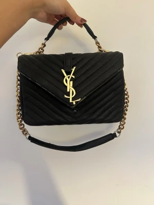 YSL väska - Ej äkta! Yves Saint Laurent väska i modellen Bolso Collége, ser äkta ut. Kort och långt band (som går att ta av). Flera fack inuti samt ett på baksidan. Väl använd så en del slitenhet på lädret och kedjan. Samt att ens knappen lossnat, därav det billiga priset. Förhoppningsvis kan nån älska väskan så mycket som jag gjorde och fixa knappen :)