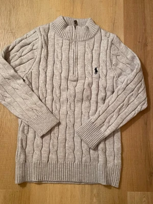 Grå kabelstickad tröja Polo Ralph Lauren - Snygg grå kabelstickad tröja från Polo Ralph Lauren med halv dragkedja och ribbade muddar. Tröjan har en klassisk passform, hög krage och den ikoniska loggan broderad på bröstet. Perfekt för lager på lager och riktigt mysig till hösten.