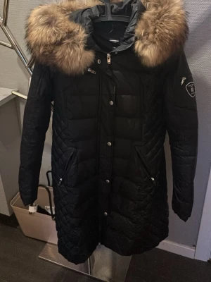 Svart Rockandblue dunparkas med pälshuva - Snygg svart lång parkas från Rockandblue med stor fluffig pälskant på huvan och quiltad design. Jackan har dragkedjor, tryckknappar och flera fickor samt en patch på ärmen. Perfekt för kalla dagar och riktigt varm.