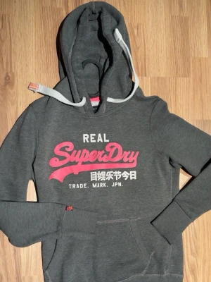 Grå Superdry hoodie med tryck - Snygg grå hoodie från Superdry med stor logga i vitt och rosa framtill. Klassisk huva med vita dragsnören och känguruficka. Tillverkad i mjukt bomullsmaterial, perfekt för en avslappnad streetwear-look. Niche