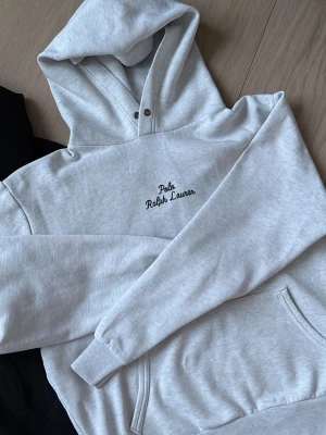 Ljusgrå hoodie från Polo Ralph Lauren - Snygg ljusgrå hoodie från Polo Ralph Lauren med broderad logga på bröstet. Klassisk känguruficka framtill och justerbar huva med snörning. Tillverkad i mjukt bomullsmaterial som är perfekt för chill dagar. Enkel och stilren design som passar till det mesta.