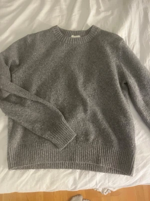 Grå stickad tröja med rund hals - Mysig grå stickad tröja med rund halsringning och ribbade muddar vid ärmslut och nederkant. Perfekt för lager-på-lager och enkel att matcha med jeans eller kjol. Klassisk och tidlös design som passar till många stilar.