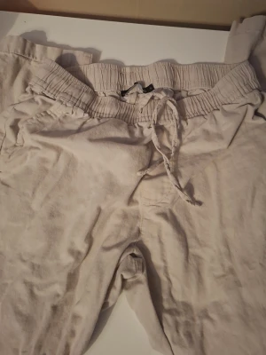 Beige linnebyxor från 157 - Säljer ett par beige linnebyxor från 157 med dragsko i midjan och resår. Byxorna har avslappnad passform och sidofickor. Perfekta för chill dagar eller när du vill ha det extra bekvämt.